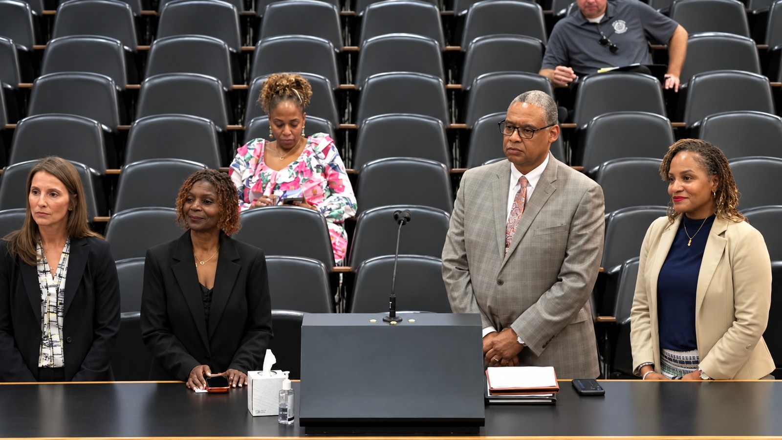 Four presenters stand at a microphone table in the Shelby County Commission chamber, with rows of chairs behind them.