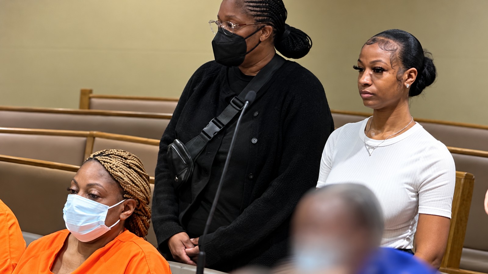 Three women appear in a courtroom, one seated in orange clothing and two standing behind her.